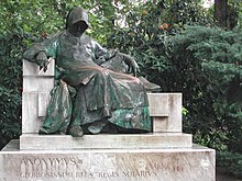 A man wearing a cowl which covers his face sits on a bench