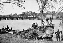 Black-and-white photo of several military men idling on a riverbank. Across the river are several large buildings