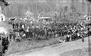 Group of indigenous people, some with instruments, pose with catholic priests. Log house, flags, and woods in background.