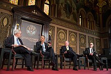 Four men in suits sit on chairs on a red stage in front of ornate gold and brown wall.