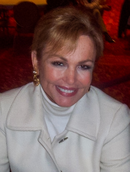 Portrait photo of a woman with blonde hair in her late 50s smiling at the camera. She is wearing a white coat and white turtleneck underneath.