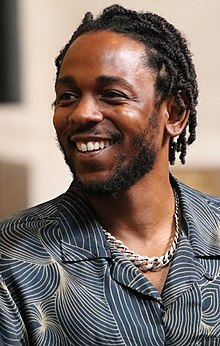A man in a dress shirt and prominent metal necklace looks camera left and smiles.