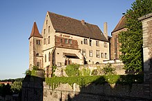 photographie en couleur montrant de loin un bâtiment rectangulaire couvert d’un enduit beige, avec une petite tourelle carrée à gauche