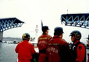 Rescue workers wearing orange uniforms on the boat approaching the bridge collapse. The two anchor trusses can be seen standing, while the middle span of the bridge is completely missing.