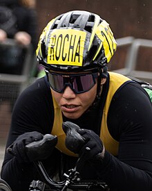 Women in black clothes with black helmet, wrap-around tinted glasses and gloves holding steering handles of a racing wheelchair. A drop of rain falls in front of her right shoulder.