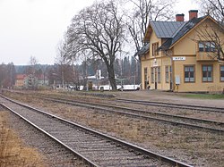 I förgrunden två rälsar och intill står ett gult hus i sekelskiftesstil med en skylt det stå Alfta på.