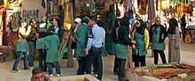 A group of young women, most in oversized green tunics and some wearing headscarves, in a bazaar. In front of them is a police officer in a blue uniform.