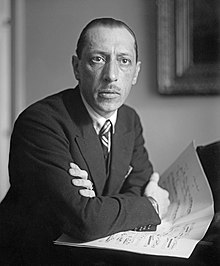 Black and white photo of Stravinsky resting his arms atop a piano, a score resting under his hands