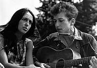 Outdoors, Joan Baez is sitting next to Bob Dylan who is playing an acoustic guitar, ca 1960s.