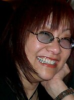 Profile picture of a bespectacled Asian woman in her early fifties. She has long red hair, and shows a toothy smile.