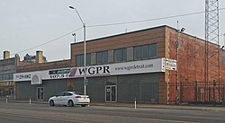 A two-story building composed of two formerly separate brick structures. The first floor has a front wall dominated by glass block. A large sign above the first floor includes the phone number and logo of WGPR radio. There are two sets of double doors and a historical marker. Next to the building is visible the bottom part of a steel lattice mast. A sign on the side of the building announces the existence of the William V. Banks Broadcast Museum and Media Center.
