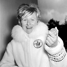 A woman with short blonde hair wearing a thick fur coat bearing a maple leaf and Olympic rings. She displays a large smile as she holds up an Olympic medal.
