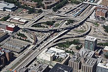 Aerial view of a major freeway interchange with several flyover ramps set within an urban neighborhood. Several ramps have exposed steel beams and other unfinished surfaces, showing signs of construction activity.