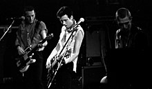 The rock band the Clash performing onstage. Three members are shown. All three have short hair. Two of the members are playing electric guitars.