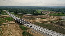 Photographie aérienne d'un échangeur en construction dans une zone plate recouverte de forêts. Alors que la chaussée de l'autoroute en construction est achevée, les bretelles ne sont que terrassées. Des véhicules de chantier et des ouvriers sont en train de travailler, tandis que des officiels peuvent être distingués, en train d'inspecter les travaux.