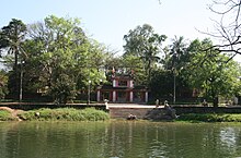 A green colored pond is at the front of a temple entrance. From the pond, there are paved stone steps up to the front of the temple, which is surrounded by a stone wall and a red triple gate with two levels and tiles. The area is surrounded by green shrubs and trees.