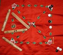 Red cloth playing surface with a crisscrossed square made of leaves and flowers, black and white tokens and four polished sticks