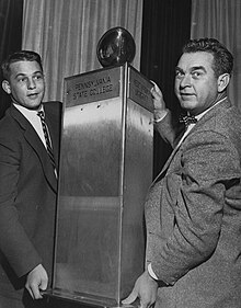 Bob Orders (left) and Art "Poppy" Lewis (right) holding the large, 3-sided steel Old Ironsides trophy. The 150 pound trophy consists of a three foot stainless steel triangular prism mounted onto a square base of unknown material. Each side of the column contains a plaque bearing the name of one of the universities, using their pre-1953 titles. Atop the trophy is a "near regulation-sized" football, presumably made of stainless steel. No further writing on the trophy or details have been documented.