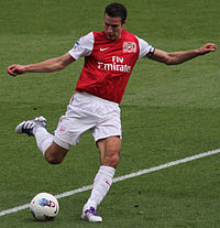 Robin van Persie, wearing a red and white football jersey and white shorts, prepares to kick a football with his right foot with both arms outstretched.