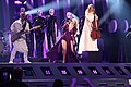 Two women—dressing purple and white dresses, respecitively—performing on a stage alongside a guitarrist; black mannequins ae seen in the background.