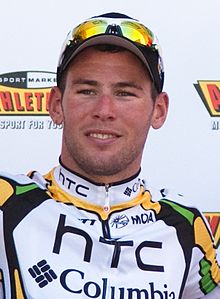 A man in his mid-twenties wearing a white and black cycling jersey with green and yellow trim, with a baseball cap and sunglasses on the top of his head. He has a thin smile on his face.