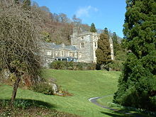 A large stone house in the middle distance, framed by trees in the foreground, and with a sloping lawn in front. The left part of the house has two storeys, while the right section is much taller, in four storeys, surmounted by battlements.