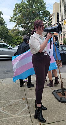 Rhea Debussy, who is a transgender woman wearing burgundy pants and a white shirt, speaking into a bullhorn and standing in front of a transgender pride flag.