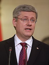 A photograph of a man with grey hair looking to the right of the viewer and wearing glasses, a black suit jacket, and a white dress shirt