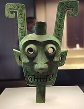 Mask showing ears, eyes, nose and mouth with teeth, with large features rising from each side of the head