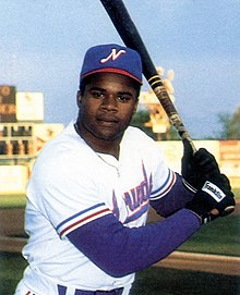 A man wearing a white baseball uniform with a blue cap on a field holding his bat, posed ready to swing