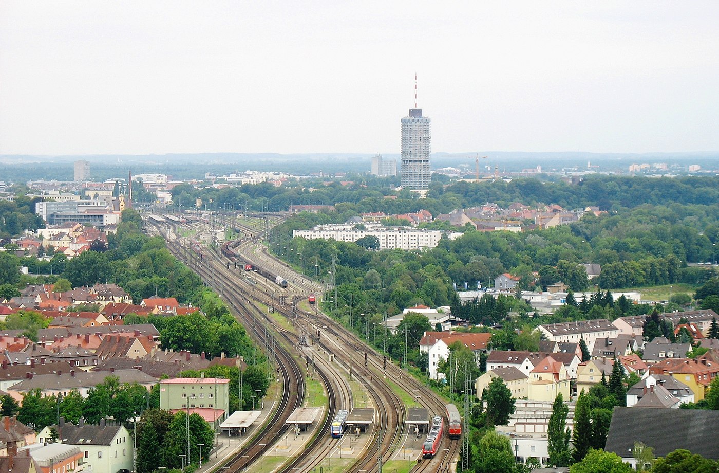 Links unten der Rand der Bahnstrecke am Augsburger Hauptbahnhof, in Bildmitte viergeschossige optisch unterteilte weiße lange Gebäude im Baumbestand: Lessing- und Schubert Hof, dahinter oben das Thelottviertel. Vorn und rechts: Wertachkanal und Bäume der Wertachaue.