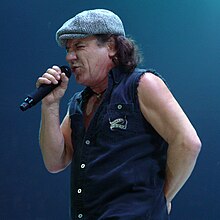 Johnson, aged 61, shown in left profile. He sings into a cordless microphone while wearing a newsboy cap and a Harley Davidson sleeveless vest.