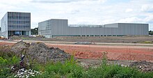 Photographie en couleur d'un bâtiment moderne en cours de construction avec au premier plan un tas de terre et une végétation de friche