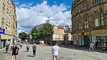 Fargate shopping precinct, Sheffield. Once a busy road, it has been pedestrianised for several decades and is Sheffield's main City Centre shopping area, home to many well known companies. The image shows classical architecture on both sides with one plan spaces in the centre, dotted with trees and the buildings on the High Street are visible beyond the trees.