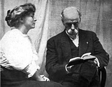 A white woman and a white man, seated. The woman, in profile, has her hair arranged in a bouffant updo, and is wearing a white blouse with puffy sleeves. The man, older, is bald, wears a mustache and a dark suit, and has his head bent to read a book.