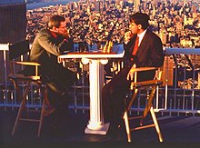 Garry Kasparov and Viswanathan Anand both sitting down at a chessboard at the beginning of a game. Both men are wearing suits and ties. Behind them is a tall railing, and a view of most of Midtown Manhattan. Anand has the White pieces, and Kasparov has the Black pieces.