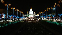 The temple grounds and Oakland California Temple at night during the Christmas season, with several plants and trees covered in Christmas lights.