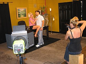 A blonde haired woman wearing a pink pantyand black miniskirt is seated onto an office desk. She seductively tugs on a man's shirt, who is standing between her thighs. In the foreground a second woman sits and photographs them, beside her is a stage light pointed towards the couple.