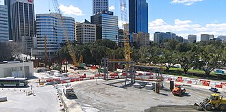 Construction site with two crawler cranes and sheet piles sticking out of the ground. Excavation of the station box has not begun yet. The city is in the background.