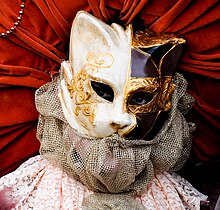 A close-up photograph of a person wearing a cat mask. The mask is colored on the left side with silver paint and on the right side with glossy black paint. Designs of shiny golden vines highlight both sides of the mask.