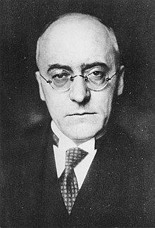 Black and white portrait of a bald, middle-aged man wearing glasses.