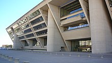 Dallas City Hall, a tall beige building with an angled front facade. The facade leans outward from the top, supported by three pillars, and is covered with rows of windows.