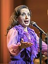 A woman with brown hair is wearing a purple blouse performing behind a microphone.