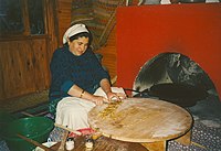 Traditional tableside preparation of gözleme in a restaurant near Antalya. A gas-powered sac griddle can be seen inside the red oven to the right, with two thin wooden rolling pins ("oklava") and pastry brushes used to prepare the dough to the lower left. The cook is seen cutting up the finished gözleme on her pastry board table to prepare for final serving.