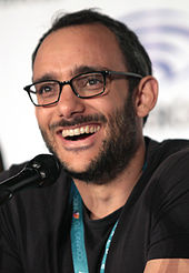 A smiling man with black hair and facial hear, wearing eyeglasses and a green lanyard, speaks into a microphone.