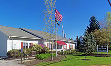 A one-story white building sided in clapboard with red shutters. The American flag flies from a pole in front, where a sign says "Red Hook Town Hall".