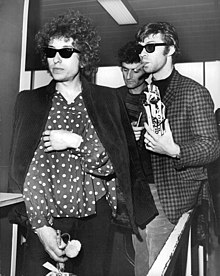 Bob Dylan, wearing sunglasses and a dark shirt with light polka dots on a dark background, with Robbie Robertson smoking a cigar and Victor Maymudes behind them.