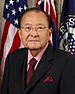 An older man wearing a suit with an American and Hawaiian flag in the background. He is seated and is staring forward toward the camera.