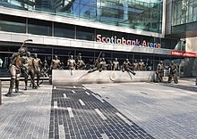 Statues of hockey players in front of a granite bench. The glass facade of Scotiabank Arena is in the background.