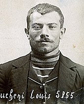 Photographie en noir et blanc d'un homme au visage légèrement souriant, portant une veste et un gilet avec un col en V qui laisse entrevoir un tricot rayé, ses cheveux sont coupés court.